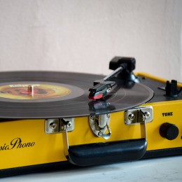 Close-up van een gele platenspeler met een zwarte vinylplaat, toonarm en toonregelaar zichtbaar.