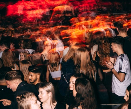 Een menigte jonge mensen in een nachtclub onder kleurrijke lichten, met beweging en energie overal.