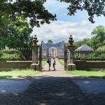 Zwei Personen stehen am Tor eines historischen Herrenhauses, umgeben von grünen Bäumen und Gärten.