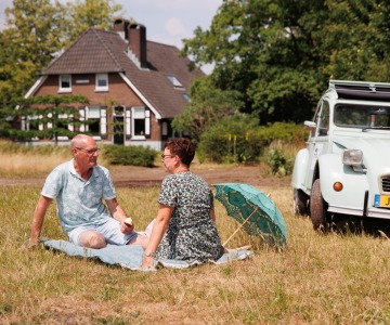 Twee mensen picknicken in een veld met een oude Citroën 2CV en een huis op de achtergrond in de zomer.