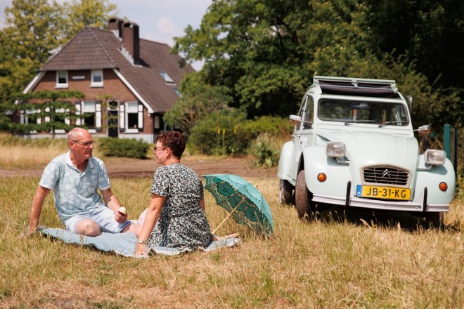 Twee mensen picknicken in een veld met een oude Citroën 2CV en een huis op de achtergrond in de zomer.