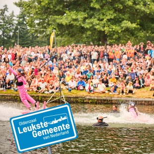 Drukke menigte kijkt naar waterwedstrijd in Oldenzaal, met deelnemer in roze pak en blauw bord op de voorgrond.