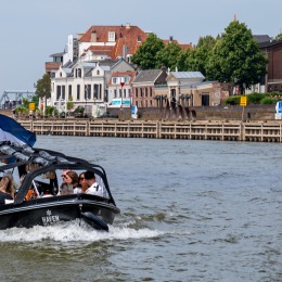 Klein zwart bootje met mensen vaart over een rivier langs huizen en boten in een Nederlandse stad.