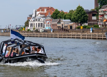 Klein zwart bootje met mensen vaart over een rivier langs huizen en boten in een Nederlandse stad.