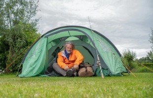Man in een oranje jas zit voor een groene tent met een tas en een vissershengel op een grasveld.