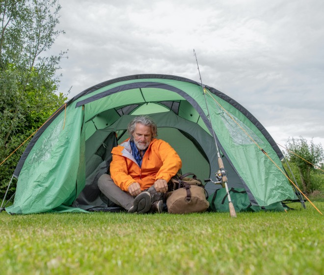 Man in een oranje jas zit voor een groene tent met een tas en een vissershengel op een grasveld.