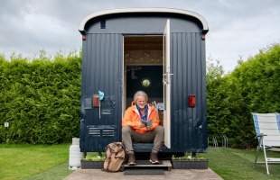 Man zit voor een tiny house, leest een boek, met rugzak naast zich op een camping omgeven door groen.