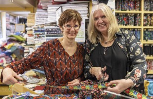 Twee vrouwen in een stoffenwinkel knippen kleurrijke stof, omringd door stapels stoffen en rollen.