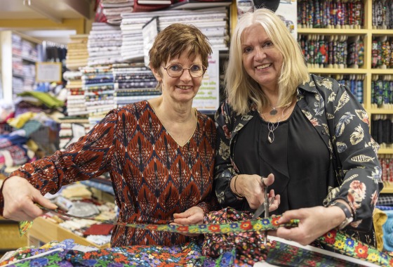Twee vrouwen in een stoffenwinkel knippen kleurrijke stof, omringd door stapels stoffen en rollen.