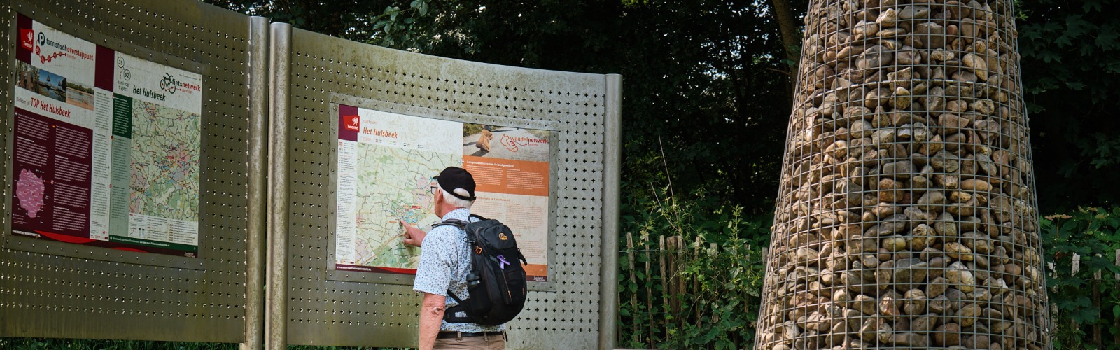 Een man met een rugzak bekijkt een informatiebord in een groen park met stenen kunstwerken.