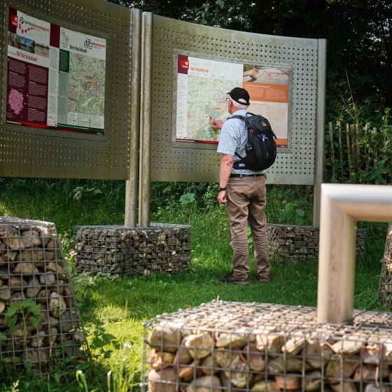 Een man met een rugzak bekijkt een informatiebord in een groen park met stenen kunstwerken.