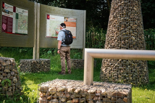 Een man met een rugzak bekijkt een informatiebord in een groen park met stenen kunstwerken.