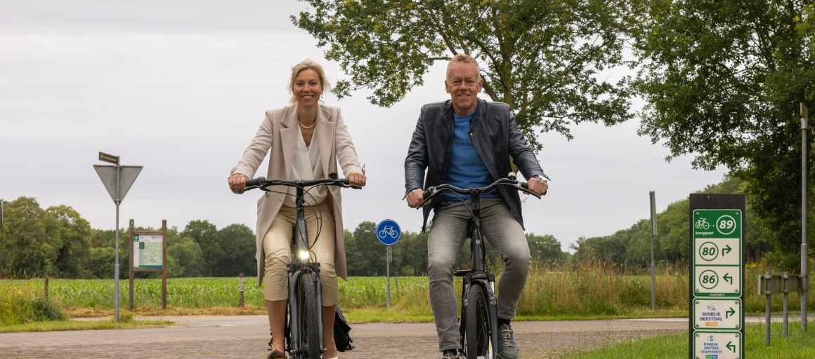 Twee mensen fietsen samen op een landweg met bewegwijzeringsborden en groen landschap op de achtergrond.