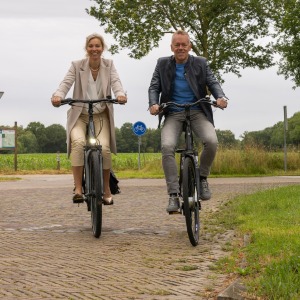 Twee mensen fietsen samen op een landweg met bewegwijzeringsborden en groen landschap op de achtergrond.