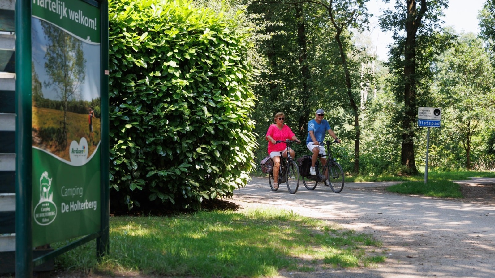 Twee mensen fietsen op een bosrijk fietspad nabij een bord van Camping De Holterberg in Nederland.