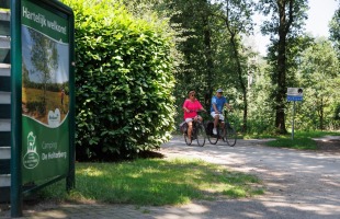 Twee mensen fietsen op een bosrijk fietspad nabij een bord van Camping De Holterberg in Nederland.