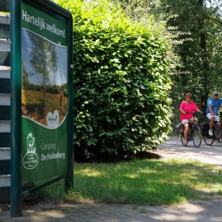 Twee mensen fietsen op een bosrijk fietspad nabij een bord van Camping De Holterberg in Nederland.