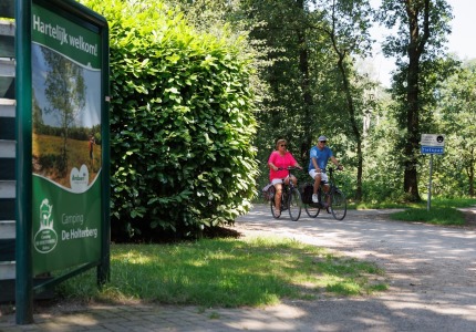 Twee mensen fietsen op een bosrijk fietspad nabij een bord van Camping De Holterberg in Nederland.