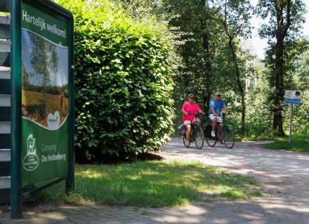 Twee mensen fietsen op een bosrijk fietspad nabij een bord van Camping De Holterberg in Nederland.