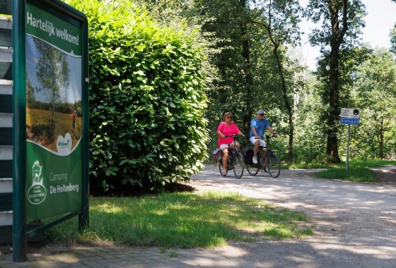 Twee mensen fietsen op een bosrijk fietspad nabij een bord van Camping De Holterberg in Nederland.