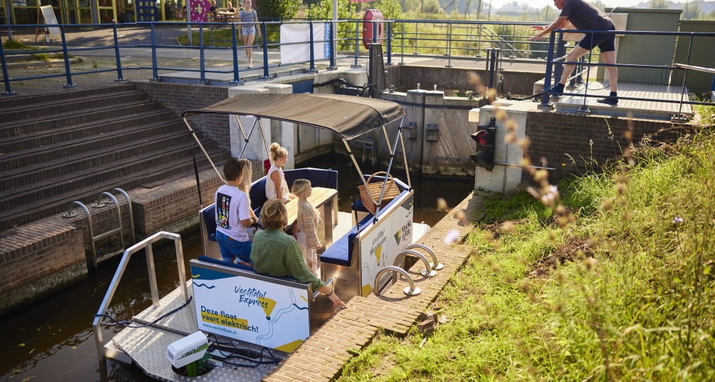 Een groep mensen zit op een elektrische boot in een sluis terwijl een man de sluis bedient.
