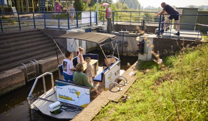 Een groep mensen zit op een elektrische boot in een sluis terwijl een man de sluis bedient.