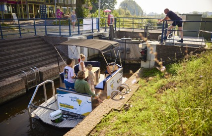 Een groep mensen zit op een elektrische boot in een sluis terwijl een man de sluis bedient.