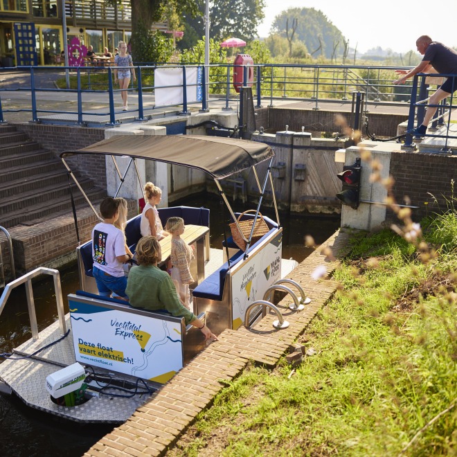 Een groep mensen zit op een elektrische boot in een sluis terwijl een man de sluis bedient.