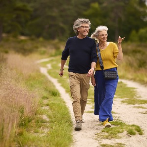 Twee oudere volwassenen wandelen vrolijk samen over een zandpad in een groen natuurlijk landschap.