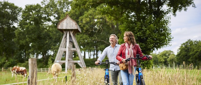Twee mensen lopen met fietsen op een landelijk pad, koeien grazen in de wei en een houten bouwwerk op de achtergrond.