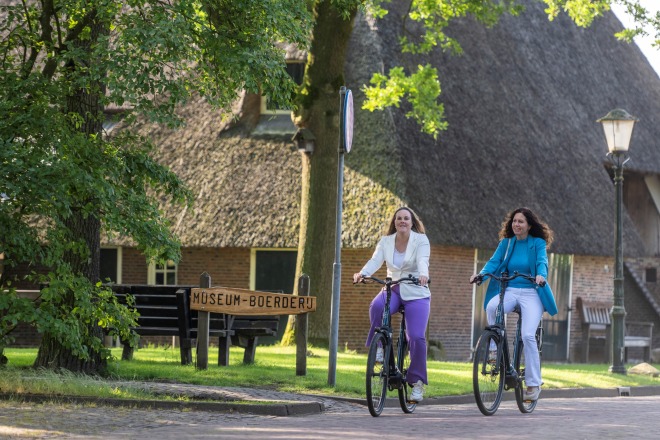 Twee vrouwen fietsen in een groene omgeving langs een bord met 'Museum-Boerderij' in Nederland.