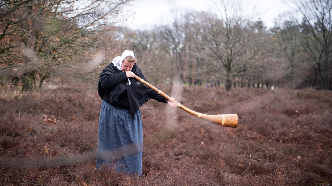 Vrouw in traditionele kleding bespeelt een houten midwinterhoorn in een herfstig, bosrijk landschap.