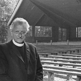 Zwart-witfoto van een geestelijke voor lege houten banken in een overdekte, openlucht kerk of amfitheater.