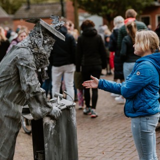 Een meisje in een blauwe jas steekt haar hand uit naar een levende standbeeldacteur in een drukke straat.