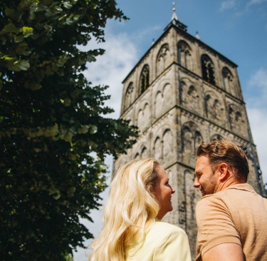 Een stel geniet samen van een zonnige dag en kijkt naar een historische kerktoren in een Nederlandse stad.
