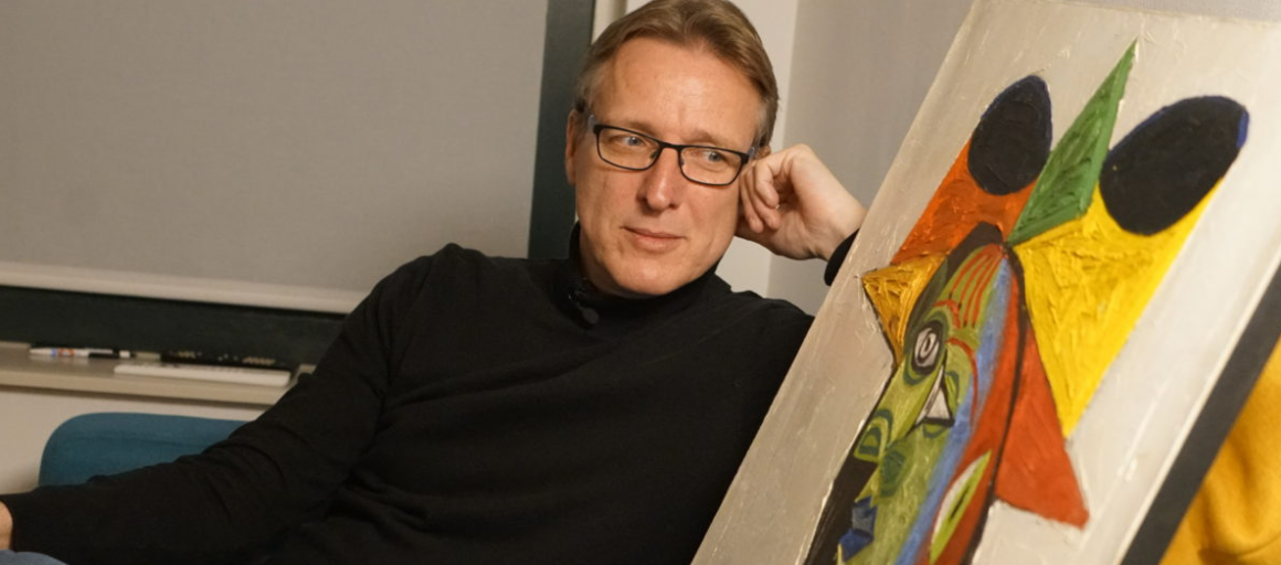 Man met bril en zwart shirt leunt achterover en kijkt naar een kleurrijk modern schilderij in de kamer.