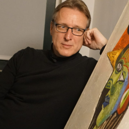 Man met bril en zwart shirt leunt achterover en kijkt naar een kleurrijk modern schilderij in de kamer.