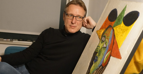 Man met bril en zwart shirt leunt achterover en kijkt naar een kleurrijk modern schilderij in de kamer.