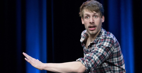 Een jonge man in een geruit overhemd gebaart op een podium met blauwe gordijnen op de achtergrond.