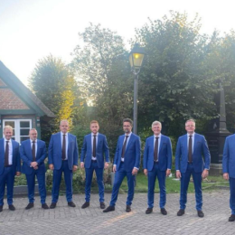 Een groep van twaalf mannen in blauwe pakken en stropdassen poseert buiten voor een huis bij zonsondergang.