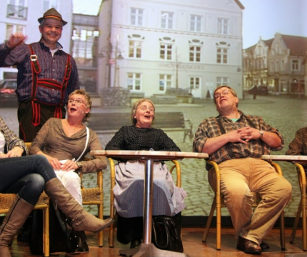 Groep mensen zit en lacht op het podium, terwijl een man in traditioneel kostuum naast hen staat.