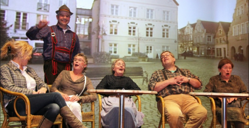Groep mensen zit en lacht op het podium, terwijl een man in traditioneel kostuum naast hen staat.