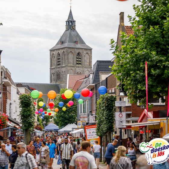Drukke straat in Nederland met kleurrijke lampionnen, kerktoren op de achtergrond en zomermarkt.