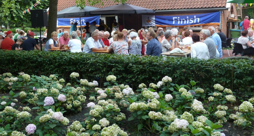 Freiluftveranstaltung mit älteren Menschen an Tischen, vor einem Haus mit 'Finish'-Banner im Garten.