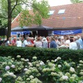 Buitenbijeenkomst met oudere mensen aan tafels, onder lombomen, voor een huis met 'Finish' spandoek.