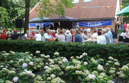 Buitenbijeenkomst met oudere mensen aan tafels, onder lombomen, voor een huis met 'Finish' spandoek.