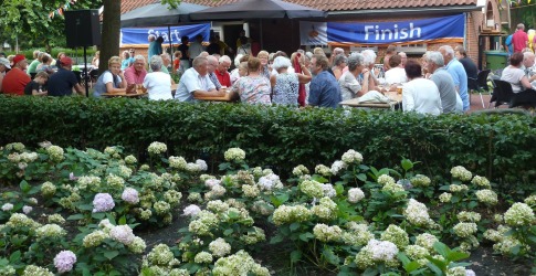 Buitenbijeenkomst met oudere mensen aan tafels, onder lombomen, voor een huis met 'Finish' spandoek.