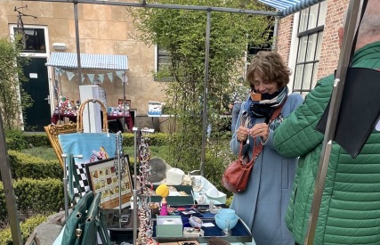 Vrouw bekijkt sieraden bij een kraampje op een buitenmarkt, onder een blauw-wit gestreepte luifel.