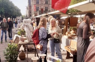 Mensen winkelen op een antiekmarkt met Franse vlag, vintage spullen en een historisch gebouw op de achtergrond.