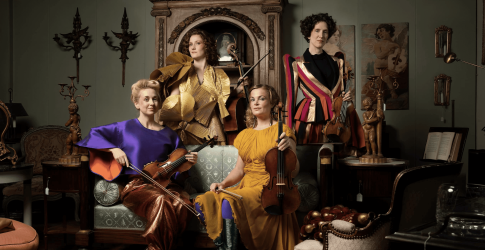 Vier vrouwen met strijkinstrumenten poseren elegant in een stijlvol ingerichte kamer met antiek.